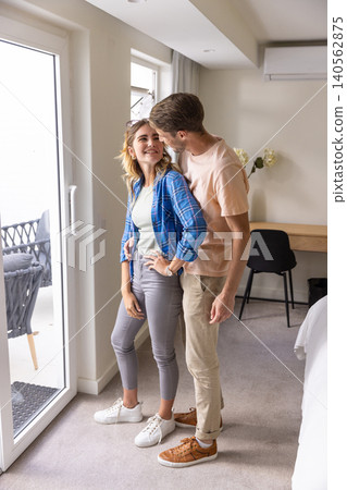 Couple embracing near sliding glass door in bedroom with balcony wicker chairs and desk flowers 140562875