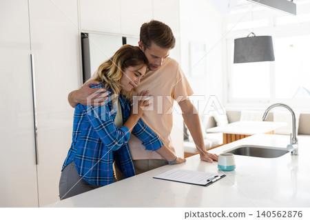 Couple embracing at modern kitchen island beside clipboard with documents, teal mug and faucet Couple embracing at modern kitchen island beside clipboard with documents, teal mug and faucet 140562876