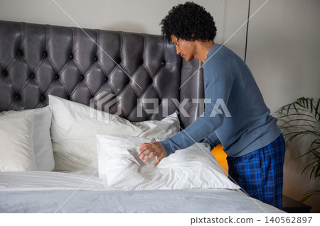 African american man fluffing pillow on bed with tufted headboard, plant in bedroom, copy space African american man fluffing pillow on bed with tufted headboard, plant in bedroom, copy space 140562897