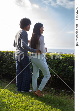 Diverse couple holding white mugs on terrace behind hedge overlooking ocean Diverse couple holding white mugs on terrace behind hedge overlooking ocean 140562898