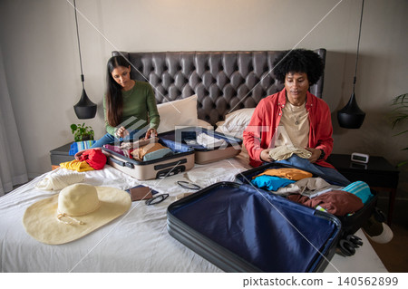 Diverse couple packing clothes into open suitcases on bed at hotel room, sun hat sunglasses Diverse couple packing clothes into open suitcases on bed at hotel room, sun hat sunglasses 140562899
