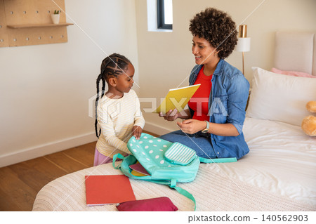 Diverse mother and daughter packing notebook and pencil case into mint-green backpack on bed Diverse mother and daughter packing notebook and pencil case into mint-green backpack on bed 140562903