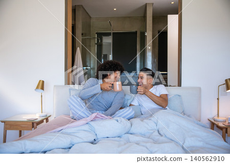 Male friends in pajamas clinking ceramic mugs while sitting on bed with duvet in bedroom Male friends in pajamas clinking ceramic mugs while sitting on bed with duvet in bedroom 140562910