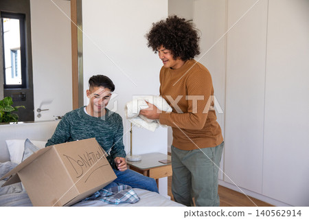 Diverse male friends packing donation box with folded clothes next to bed in bedroom Diverse male friends packing donation box with folded clothes next to bed in bedroom 140562914