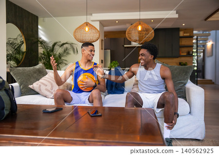 Diverse male friends sitting on sofa high-fiving, holding basketball and smartphone in living room Diverse male friends sitting on sofa high-fiving, holding basketball and smartphone in living room 140562925