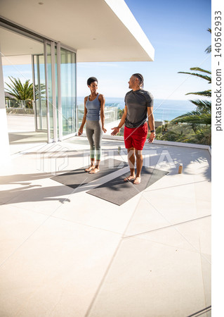 Diverse couple performing yoga on mats at modern balcony overlooking ocean with water bottles 140562933
