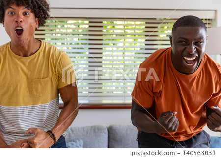 Diverse male friends cheering and leaning forward in living room near couch with decorative pillows Diverse male friends cheering and leaning forward in living room near couch with decorative pillows 140563031