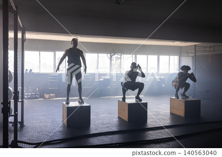 Diverse male friends squatting on wooden plyometric boxes in gym with dumbbells and weight plates Diverse male friends squatting on wooden plyometric boxes in gym with dumbbells and weight plates 140563040