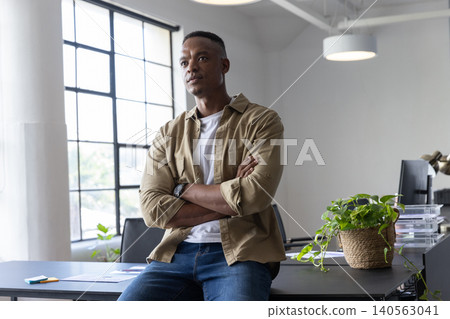 African american man sitting on desk, gazing toward window in office with monitor, plant 140563041