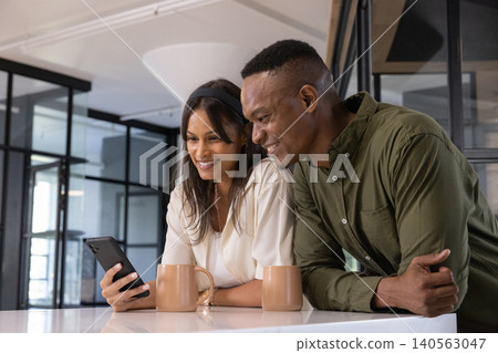 Diverse coworkers leaning on white countertop in kitchenette, checking smartphone near mugs Diverse coworkers leaning on white countertop in kitchenette, checking smartphone near mugs 140563047