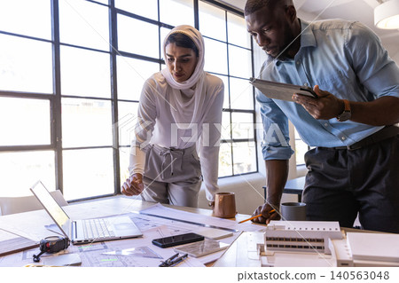 Diverse coworkers collaborating at office wearing business attire reviewing blueprints with tablet Diverse coworkers collaborating at office wearing business attire reviewing blueprints with tablet 140563048