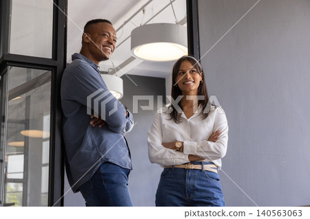 Diverse coworkers leaning with crossed arms near glass partition, pendant lamps at modern office Diverse coworkers leaning with crossed arms near glass partition, pendant lamps at modern office 140563063