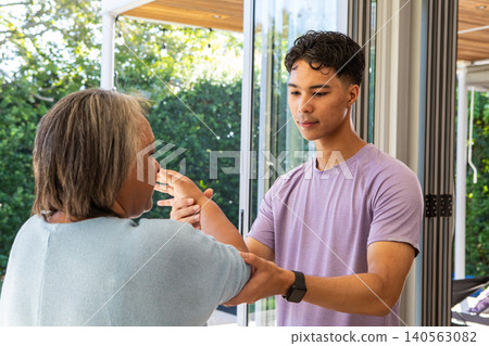 Senior Hispanic woman stretching arms as Asian man guiding movement on home patio with glass doors Senior Hispanic woman stretching arms as Asian man guiding movement on home patio with glass doors 140563082