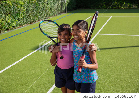 Asian female children friends sharing joyful moment holding rackets on outdoor court with lines Asian female children friends sharing joyful moment holding rackets on outdoor court with lines 140563107