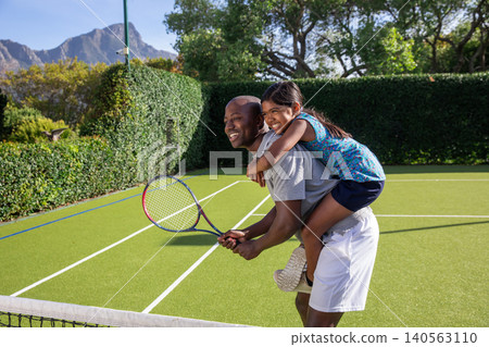 Asian girl riding piggyback on African American father gripping tennis racket by net on court Asian girl riding piggyback on African American father gripping tennis racket by net on court 140563110