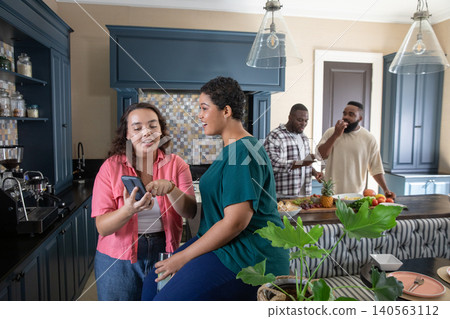 Diverse friends leaning over smartphone at kitchen island with cloth and fruit bowl Diverse friends leaning over smartphone at kitchen island with cloth and fruit bowl 140563112