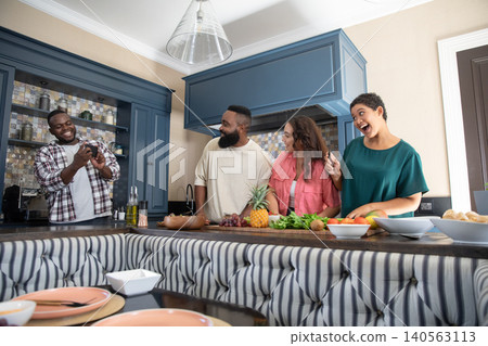 Diverse friends standing behind kitchen island, showing smartphone screen near pineapple and grapes 140563113