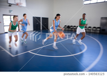 Diverse female players dribbling basketball under hoop on 3-point arc in gym with folding chairs Diverse female players dribbling basketball under hoop on 3-point arc in gym with folding chairs 140563123