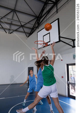 Diverse female athletes leaping toward hoop while defenders raising arms on basketball court Diverse female athletes leaping toward hoop while defenders raising arms on basketball court 140563126
