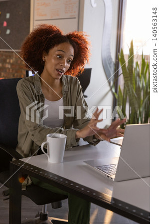 African american woman gesturing while speaking at desk in office with laptop and coffee mug 140563148