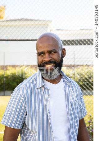 Senior African American man smiling and looking right standing at chain-link fence near grassy lawn 140563149