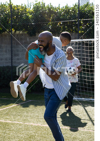 African American family lifting child near goal net and fence on artificial turf with soccer ball African American family lifting child near goal net and fence on artificial turf with soccer ball 140563150