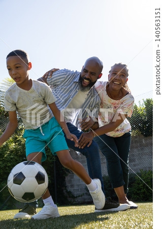 African American family playing with soccer ball in backyard near chain-link fence and hedge 140563151
