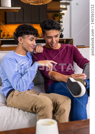 Male friends leaning toward each other viewing tablet on gray sofa in living room Male friends leaning toward each other viewing tablet on gray sofa in living room 140563152