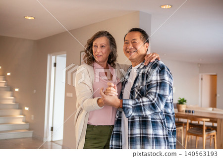 Diverse senior couple dancing slowly in open-plan living area with lit staircase, dining table 140563193