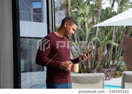African American man standing on modern poolside terrace holding coffee mug and checking smartphone African American man standing on modern poolside terrace holding coffee mug and checking smartphone 140563202