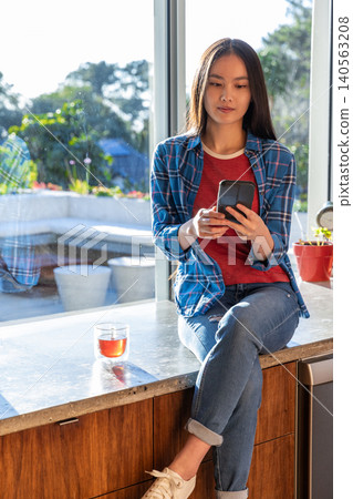 Asian woman sitting on marble counter by window holding smartphone with glass tea mug 140563208