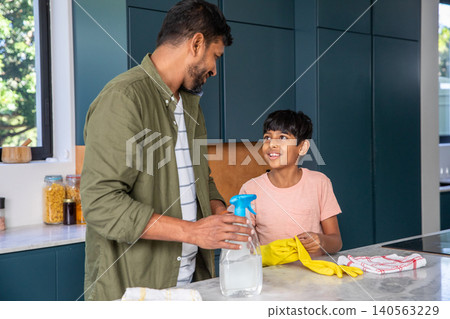 Indian father with son standing at countertop in home kitchen spraying cleaner while wearing gloves Indian father with son standing at countertop in home kitchen spraying cleaner while wearing gloves 140563229