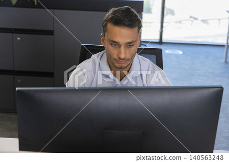 Hispanic male working at white desk in office using large monitor with mesh chair Hispanic male working at white desk in office using large monitor with mesh chair 140563248
