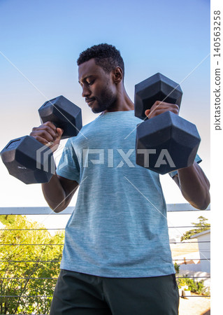 African american man in blue shirt and black shorts, gripping hex dumbbells on balcony railing African american man in blue shirt and black shorts, gripping hex dumbbells on balcony railing 140563258