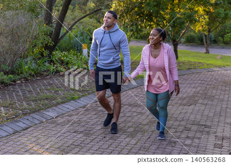 Diverse couple walking hand in hand along path in park among shrubs under sunlit trees Diverse couple walking hand in hand along path in park among shrubs under sunlit trees 140563268