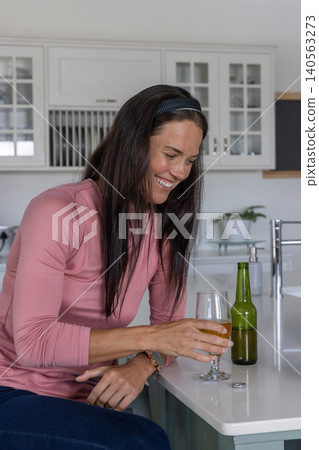 Woman in her 30s sitting in kitchen holding beer with green beer bottle on countertop 140563273