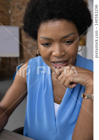 African American woman in blouse leaning on hand reviewing notes at desk with whiteboard in office African American woman in blouse leaning on hand reviewing notes at desk with whiteboard in office 140563296