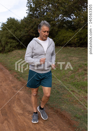 Senior man jogging on dirt trail wearing hoodie and athletic shorts with running shoes Senior man jogging on dirt trail wearing hoodie and athletic shorts with running shoes 140563305