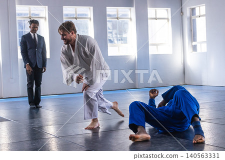 Male practitioner in white gi rising to feet on dojo mats with instructor observation Male practitioner in white gi rising to feet on dojo mats with instructor observation 140563331