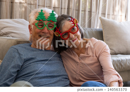 Senior couple leaning heads together and smiling on sofa at home wearing novelty Christmas glasses Senior couple leaning heads together and smiling on sofa at home wearing novelty Christmas glasses 140563370
