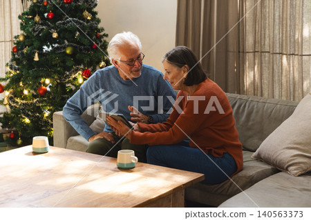 Senior couple sitting on gray sofa using tablet in living room near Christmas tree and mugs Senior couple sitting on gray sofa using tablet in living room near Christmas tree and mugs 140563373