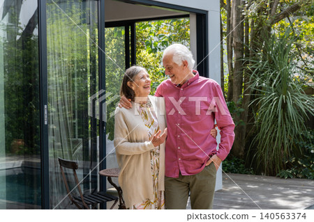 Senior couple walking arm in arm on wooden deck patio by small round table outside home Senior couple walking arm in arm on wooden deck patio by small round table outside home 140563374