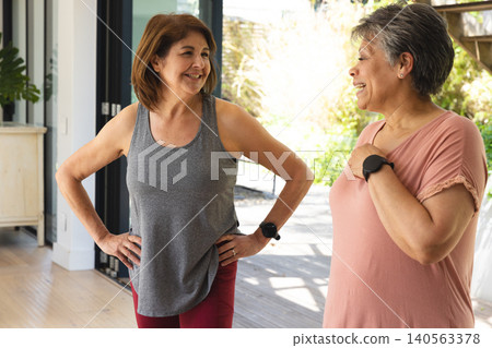 Senior women friends standing and talking at home with fitness trackers near sliding glass doors Senior women friends standing and talking at home with fitness trackers near sliding glass doors 140563378