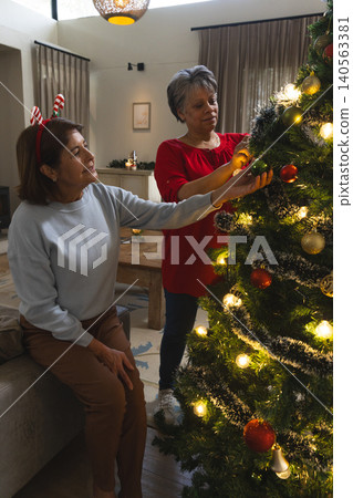 Senior female friends decorating Christmas tree at home by coffee table wearing antler headband Senior female friends decorating Christmas tree at home by coffee table wearing antler headband 140563381