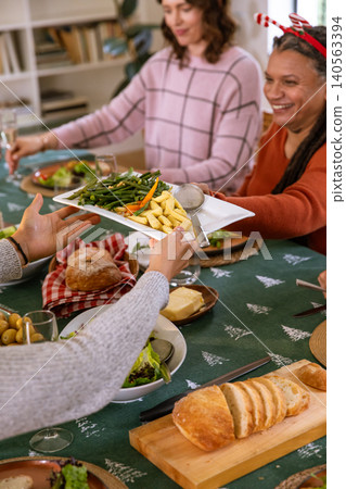 Diverse family in festive sweaters passing platter of green beans, potatoes around dining table Diverse family in festive sweaters passing platter of green beans, potatoes around dining table 140563394