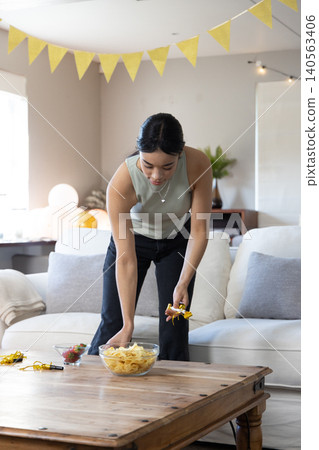 Asian woman arranging glass bowl with chips, party blowers on coffee table in living room Asian woman arranging glass bowl with chips, party blowers on coffee table in living room 140563406