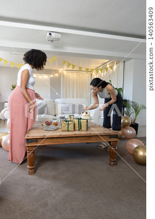 Diverse female friends preparing small cake on wooden coffee table in living room with gift boxes Diverse female friends preparing small cake on wooden coffee table in living room with gift boxes 140563409