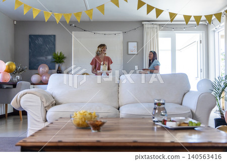 Female friends with cake and cups while standing behind sofa at home with balloons and bunting Female friends with cake and cups while standing behind sofa at home with balloons and bunting 140563416