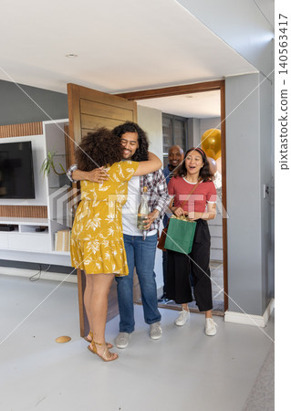Diverse friends hugging couple at home entrance, holding wine bottle, gold balloons, gift bag Diverse friends hugging couple at home entrance, holding wine bottle, gold balloons, gift bag 140563417
