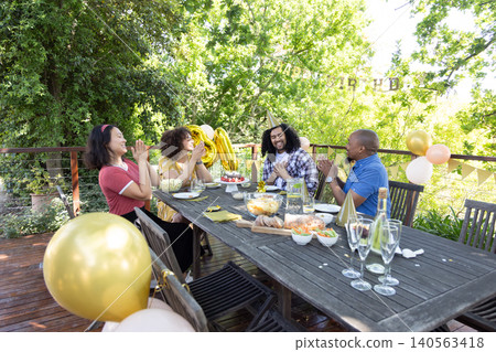 Diverse friends clapping and laughing around cake charcuterie board, balloons on backyard deck 140563418